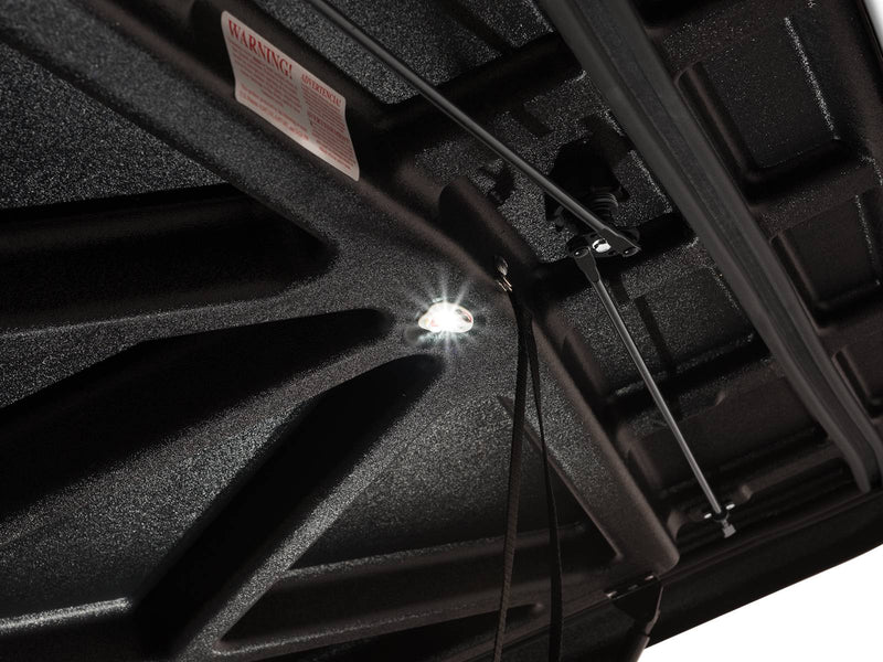 Close-up of the small round LED puck light lit against the black textured X-brace molding under the raised SE lid, with the warning decal alongside.