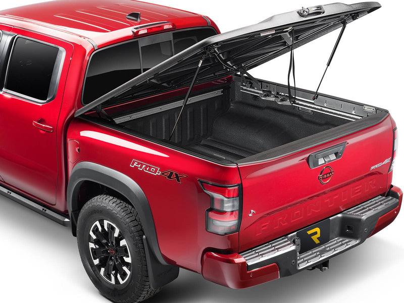 UnderCover Elite one-piece tonneau cover raised on its two hydraulic struts to about 45 degrees, rear three-quarter view, the molded X-brace underside and the empty spray-lined bed floor both visible.