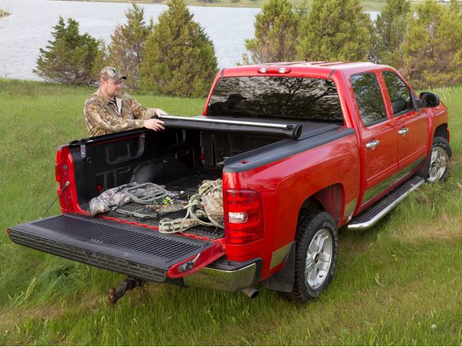 Man in camouflage rolling the cover toward the cab of a red pickup, tailgate down, gear in the bed