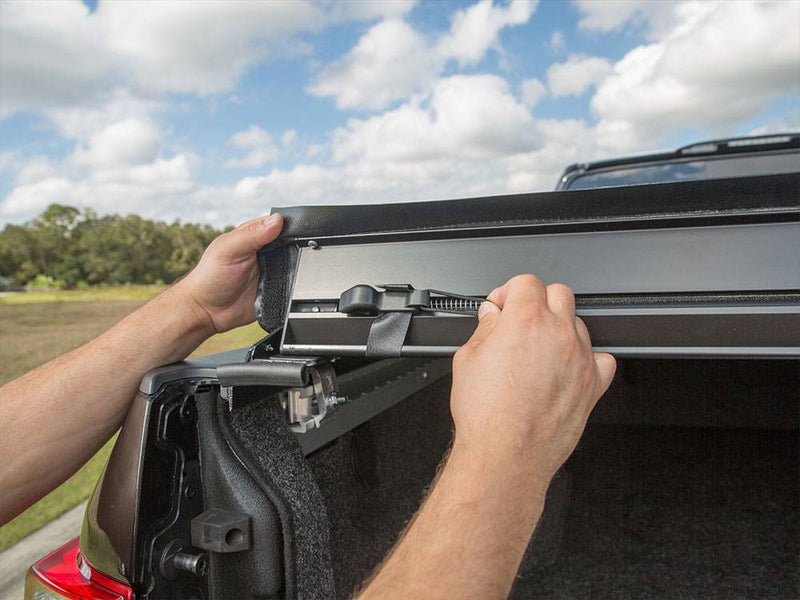 Hands lifting the rear edge of the cover to show the tension bar and latch at the tailgate