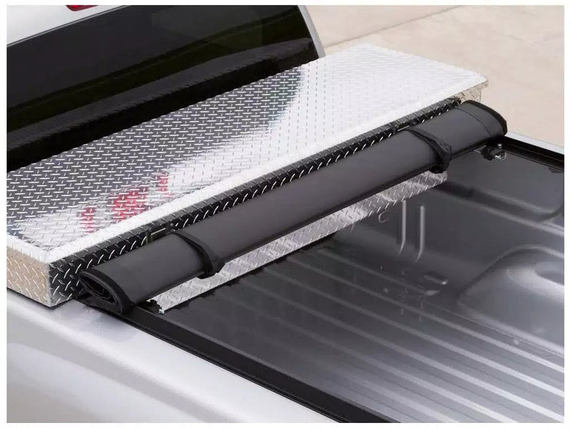 Close-up of the Access Toolbox Edition roll-up tonneau cover: close-up of a black roll-up cover rolled and strapped against an aluminum diamond-plate bed toolbox (truck-bed toolbox shown, not included)