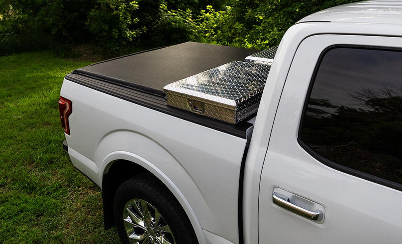 Access Toolbox Edition roll-up tonneau cover on a pickup bed: three-quarter side view of a white pickup on grass, black roll-up cover closed behind an aluminum diamond-plate bed toolbox (truck-bed toolbox shown, not included)