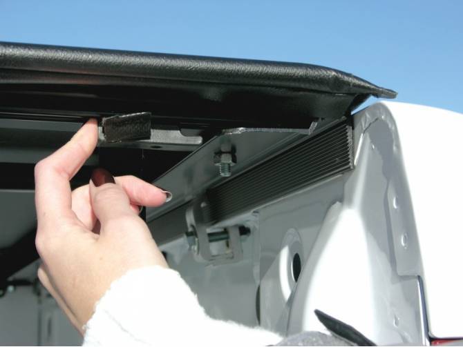 Hand pulling the rear latch release tab under the lifted rear edge of the cover