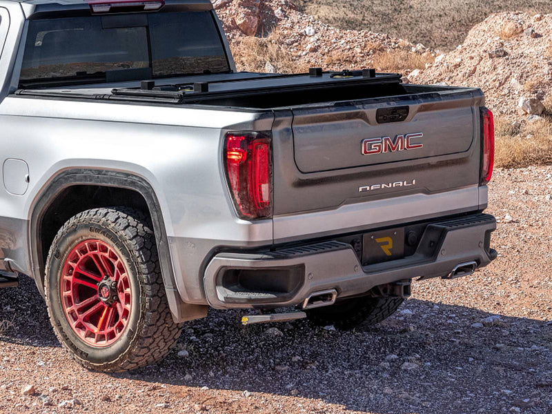 Rear corner of a silver pickup with red wheels on gravel, the bed step folded under the bumper