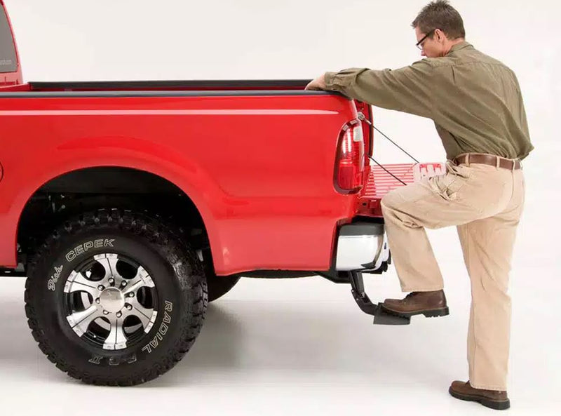A man stands on the BedStep to reach over the bed side of a red heavy-duty pickup, studio background, second angle