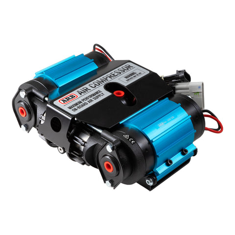 ARB CKMTA12 twin motor on-board 12V air compressor, two blue anodized motor barrels flanking a black manifold cover printed ARB AIR COMPRESSOR MAXIMUM PERFORMANCE ON-BOARD AIR SUPPLY, with two black cylinder heads, red inlet grommets and a wiring harness