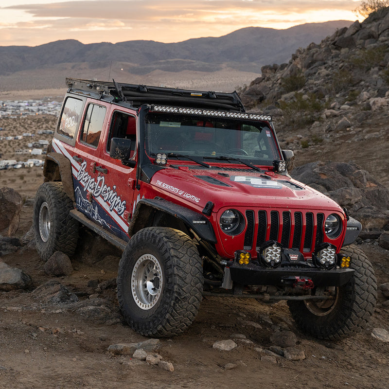 Baja Designs OnX6+ 50" Roof Mount Light Kit installed on a Jeep Wrangler JL: A red Jeep Wrangler with Baja Designs livery crawls rocky desert terrain with a long LED light bar mounted above the windshield