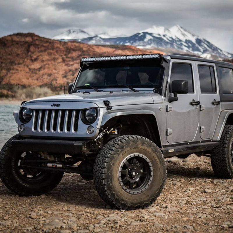 Baja Designs OnX6+ 50" Roof Mount Light Kit installed on a Jeep Wrangler JK: silver four-door Jeep Wrangler JK on rocky ground with a light bar mounted over the windshield and mountains behind