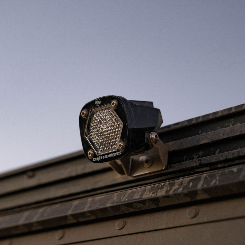 Baja Designs S1 Black LED Auxiliary Light Pod on a vehicle: A black S1 light pod with a clear dimpled lens mounted on a metal rack rail at dusk