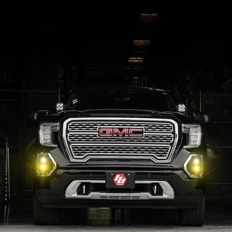 Baja Designs S1 Dual Fog Pocket Light Kit installed on a GMC Sierra 1500: black GMC Sierra viewed head-on in a dark garage with amber S1 fog lights glowing and pods on the A-pillars