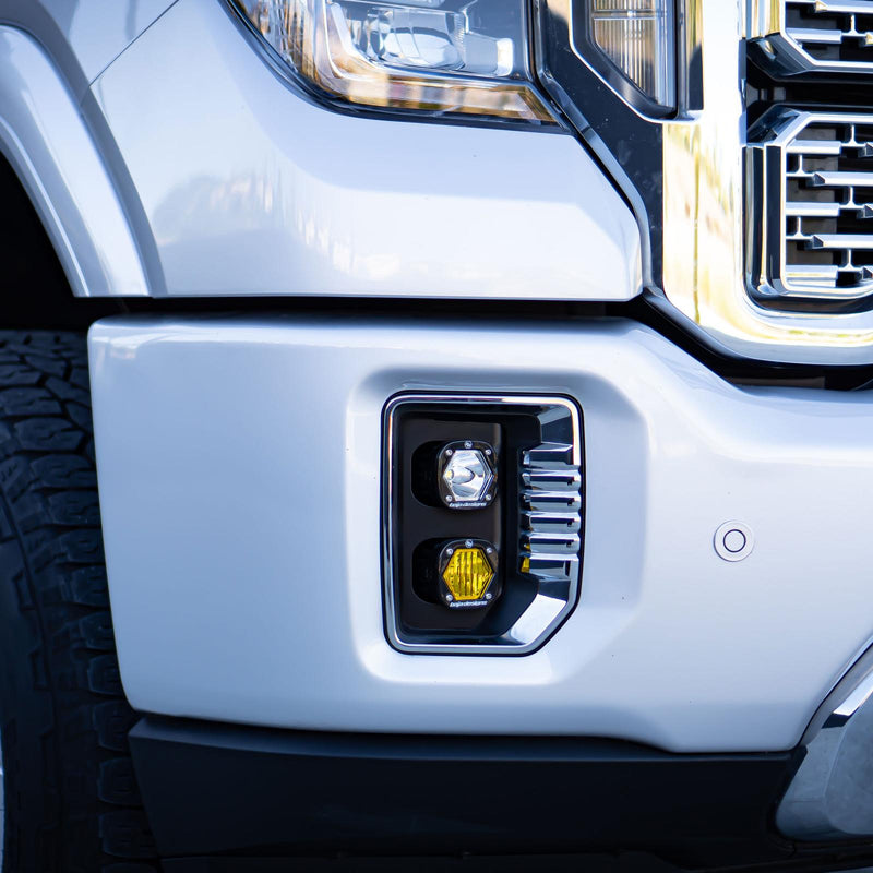 Baja Designs S1 Dual Fog Pocket Light Kit on a GMC Sierra HD: close view of a white truck front bumper with a dual pod fog pocket light, clear pod above an amber pod, below the headlight and chrome grille