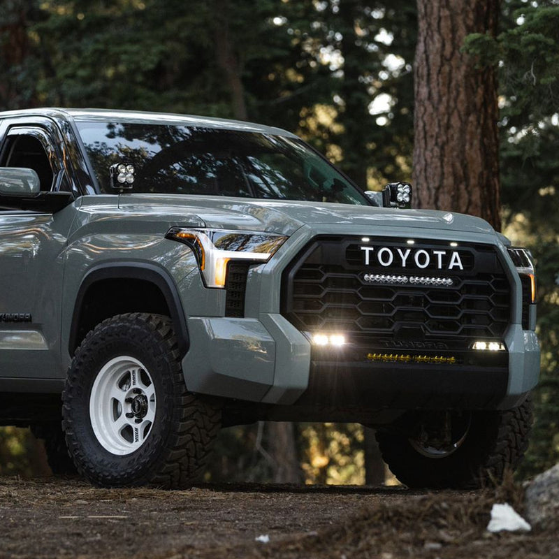 Baja Designs S2 SAE OEM Fog Light Replacement Kit installed on a Toyota Tundra: gray Toyota Tundra parked in a pine forest with its lower fog lights and grille light glowing