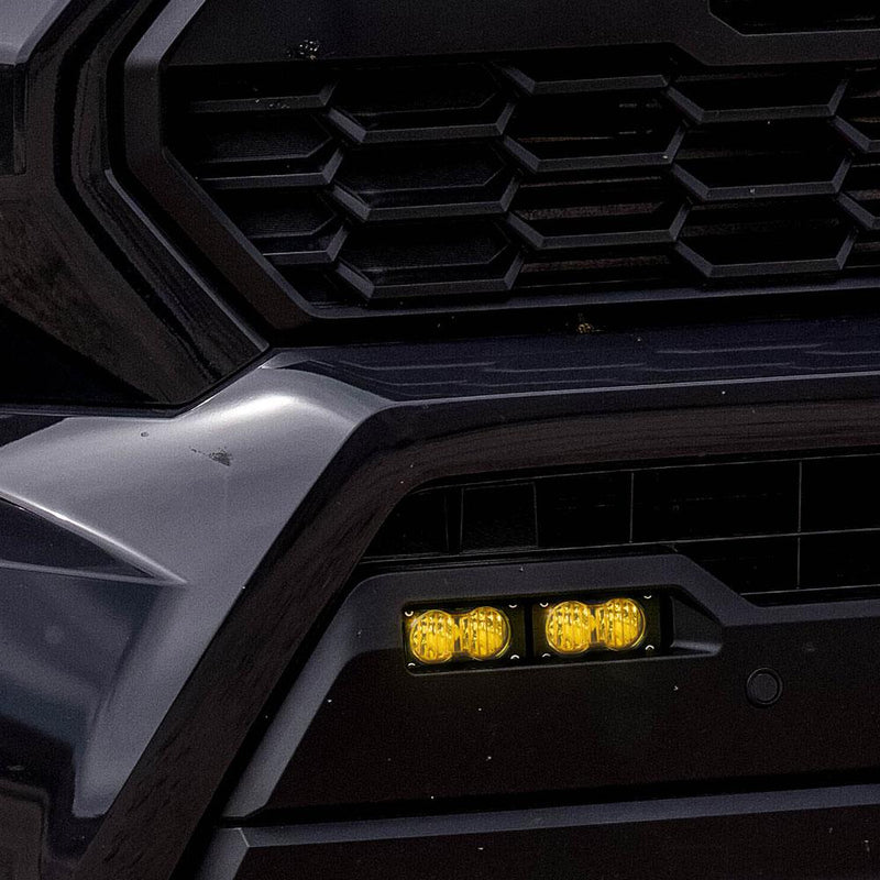 Baja Designs S2 Sport Dual Fog Pocket Light Kit on a Toyota Tacoma: close view of two lit amber S2 pods in the lower fog pocket below a dark Toyota hexagon grille