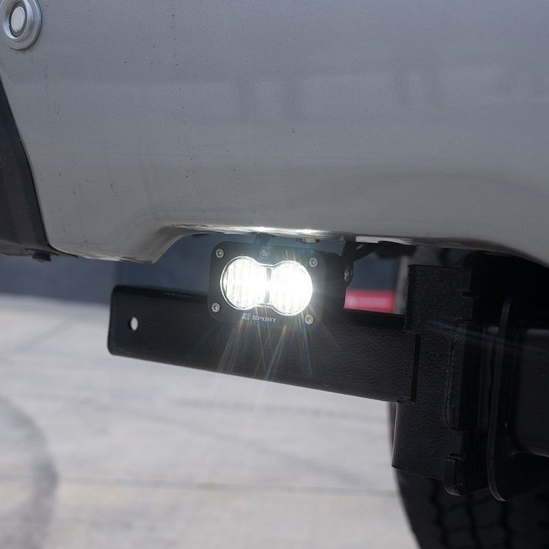 Baja Designs S2 Sport Dual Reverse Light Kit on a vehicle: A lit S2 Sport LED pod mounted next to the receiver hitch under a silver truck rear bumper