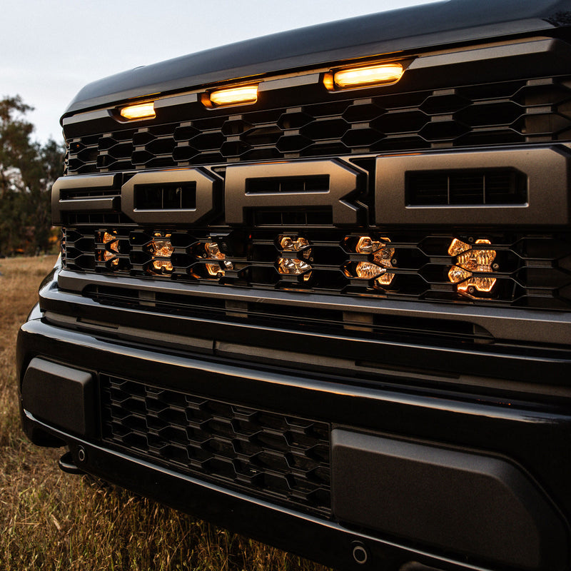 Baja Designs Squadron 2.0 Pro Behind-the-Grille Light Kit on a Ford F-150 Raptor: close view of a black Ford Raptor grille with warm glowing pods behind the slots beneath the FORD lettering