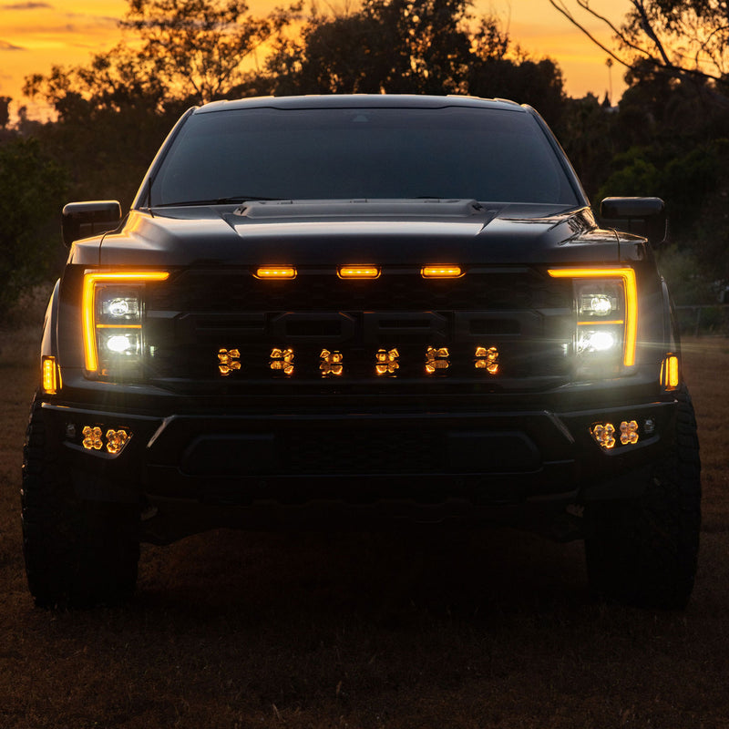 Baja Designs Squadron 2.0 Sport Behind-the-Grille Light Kit installed on a Ford F-150 Raptor: front view of a black Ford Raptor at dusk with six lit pods behind the grille and lit fog lights