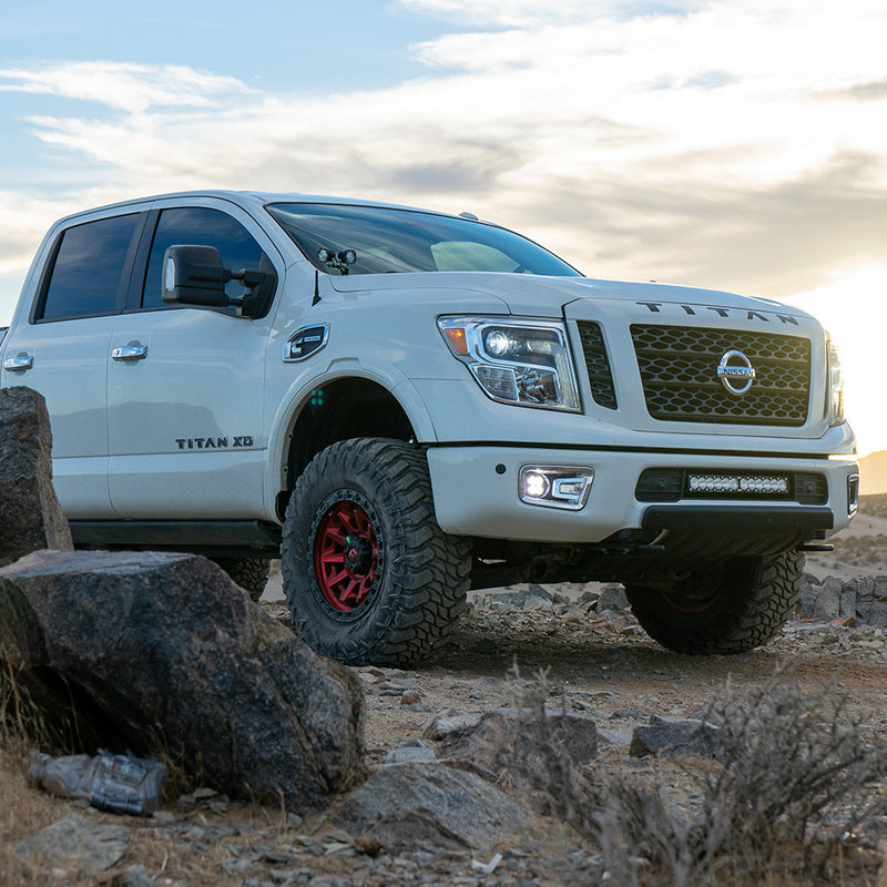 Baja Designs Squadron-R Pro Fog Pocket Light Kit installed on a Nissan Titan XD: A white Nissan Titan XD parked among desert rocks with clear fog lights glowing and a lower bumper light bar