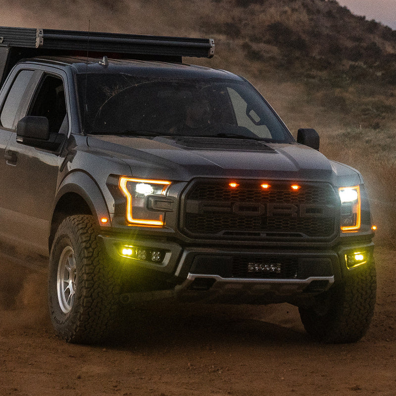 Baja Designs Squadron/S2 SAE Pro Fog Pocket Light Kit installed on a Ford F-150 Raptor: A gray Ford Raptor driving on dirt with amber fog pocket lights glowing in the bumper