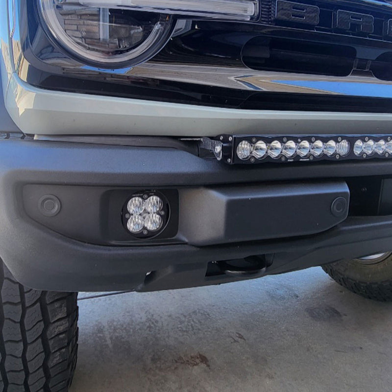 Baja Designs Squadron Sport Fog Pocket Light Kit on a Ford Bronco: close view of a clear Squadron pod mounted in the fog pocket of a Ford Bronco bumper with a light bar above it and BRONCO lettering on the grille