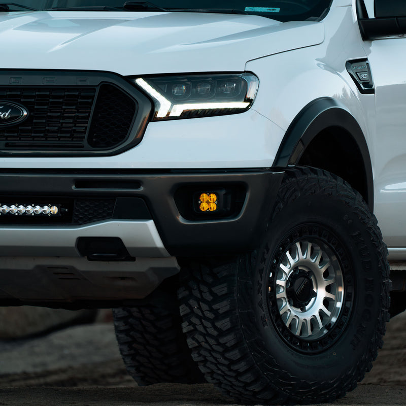 Baja Designs Squadron Sport Fog Pocket Light Kit installed on a Ford Ranger: white Ford Ranger front three-quarter view with an amber fog pocket light lit in the bumper and a light bar in the lower grille; title does not state lens color