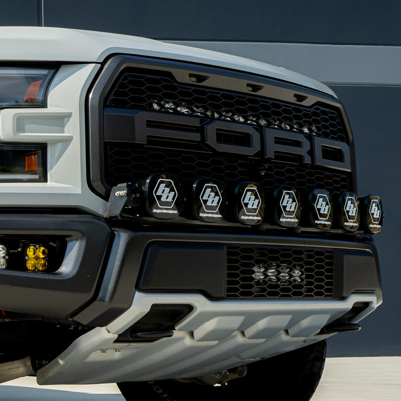 Baja Designs XL Linkable Bumper Light Kit on a Ford F-150 Raptor: close front view of a white Ford Raptor grille with XL pods wearing Baja logo covers mounted below the FORD lettering, and an amber fog pod
