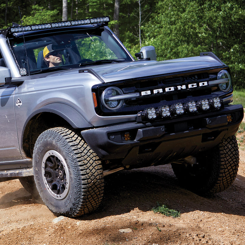 Baja Designs XL Linkable Bumper Light Kit on a Ford Bronco: front corner of a silver Ford Bronco on a dirt trail with six linkable LED pods mounted across the front bumper