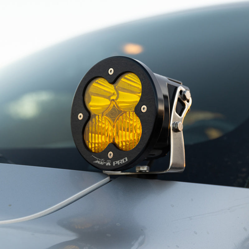Baja Designs XL-R Pro LED Auxiliary Light Pod Pair on a vehicle: A round black XL-R Pro pod with an amber lens mounted near the hood edge of a gray vehicle, close up