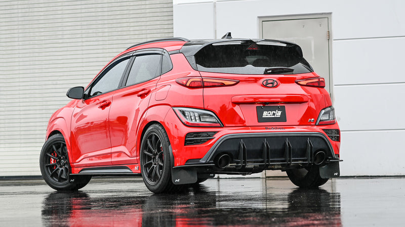 Borla ATAK Cat-Back Exhaust System installed on a Hyundai Kona N: red Hyundai Kona N hatchback seen from the rear quarter on a wet lot, with a Borla plate and rear diffuser