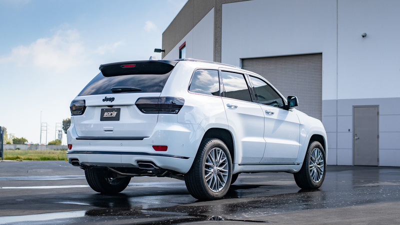 Borla S-Type Cat-Back Exhaust System installed on a Jeep Grand Cherokee: white Jeep Grand Cherokee seen from the rear quarter beside a building, with a Borla plate and exhaust tips at each bumper corner