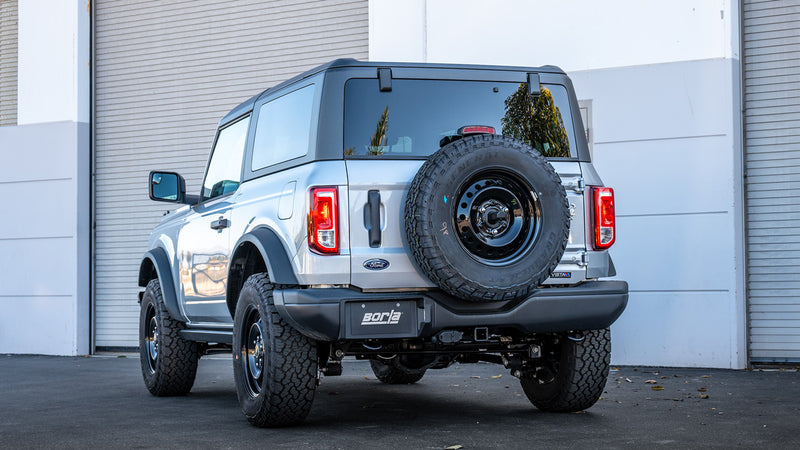 Borla Touring Cat-Back Exhaust System installed on a Ford Bronco: A silver Ford Bronco with a rear-mounted spare tire parked by a white garage, seen from directly behind