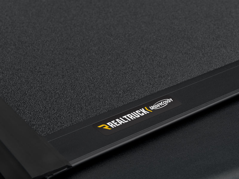 Close-up of the RealTruck UnderCover nameplate badge on the textured matte-black panel surface of the ArmorFlex cover.
