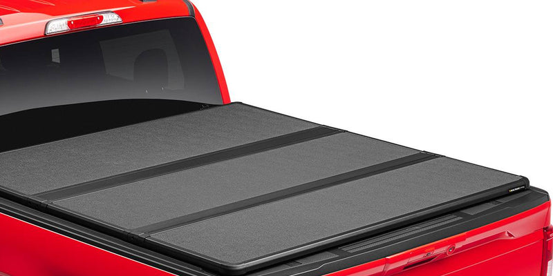 Closed hard tri-fold tonneau cover with three matte panels over a red pickup bed, rear three-quarter close-up