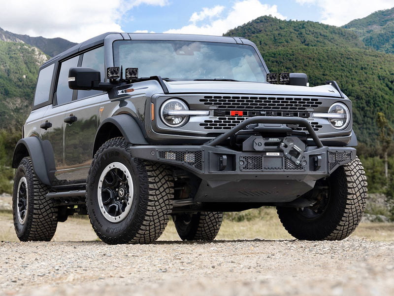 Go Rhino Rockline: gray 4-door SUV front three-quarter on gravel with a textured black winch bumper and overrider hoop (shown with winch, cube lights, A-pillar cube lights, not included)