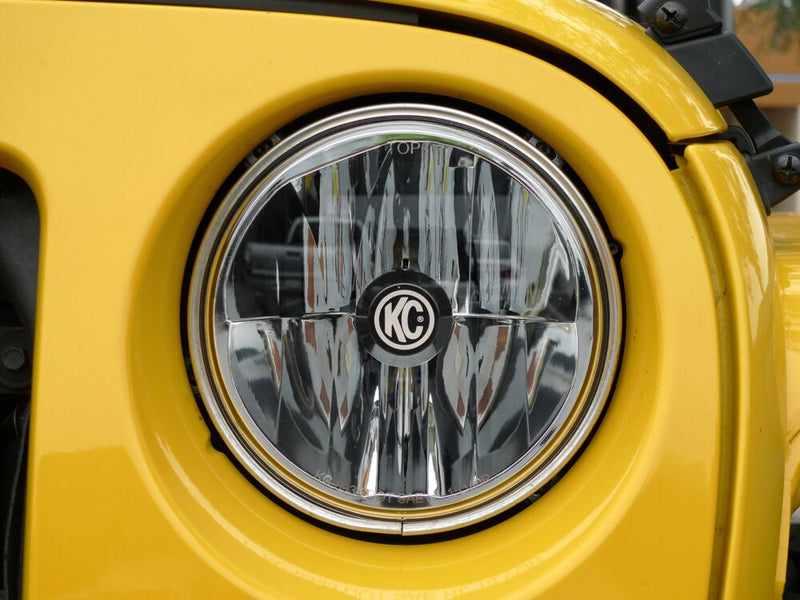 KC HiLiTES 7" Gravity LED DOT Headlight (H4 plug) installed on a Jeep Wrangler: close-up of a round LED headlight with KC center badge installed in a yellow Jeep grille