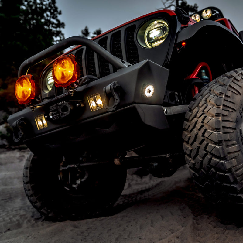KC HiLiTES Cyclone V2 LED Light on a Jeep Wrangler JL: low angle of a red Jeep front end on sand with a small round white light glowing in the bumper corner and amber round lights on the bar