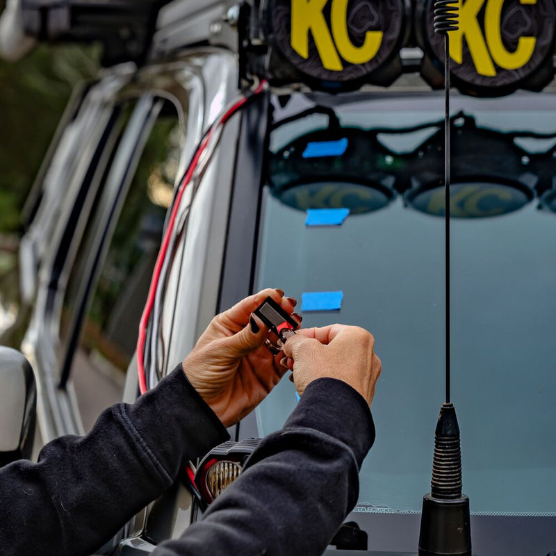 KC HiLiTES Universal Wire Hider with KClips on a Toyota: hands pressing a wire hider clip onto the windshield pillar of a silver truck, with KC lights on the roof rack and a CB antenna