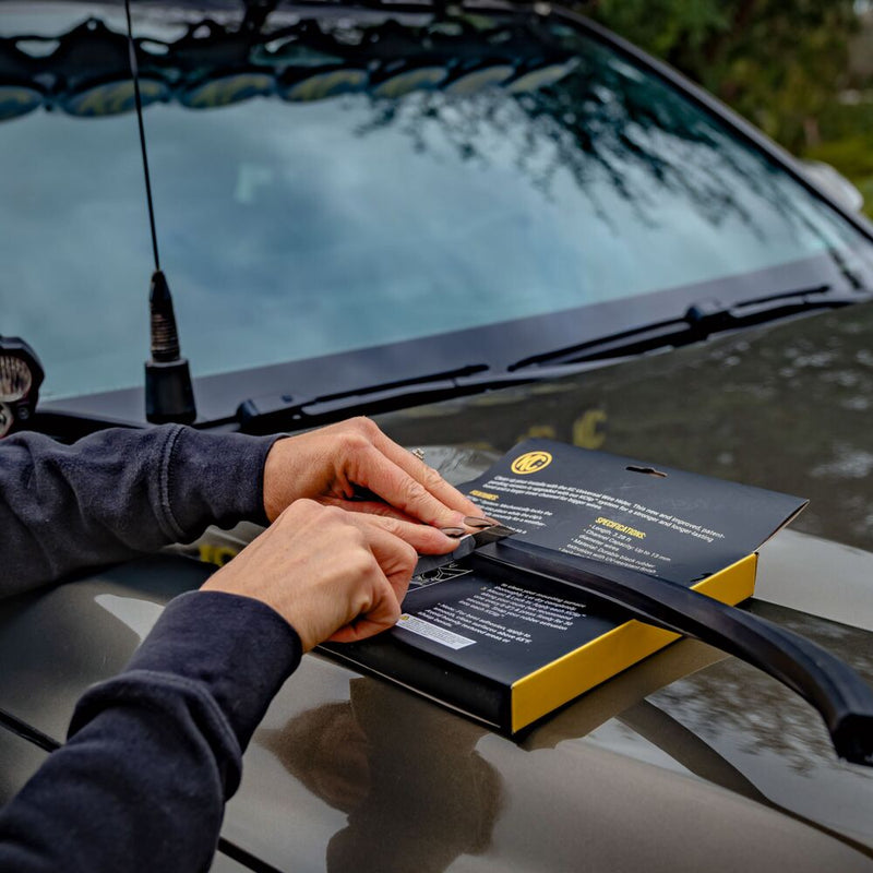 KC HiLiTES Universal Wire Hider with KClips on a Toyota: hands pulling a black wire hider strip from its KC retail package resting on the hood of a gray truck