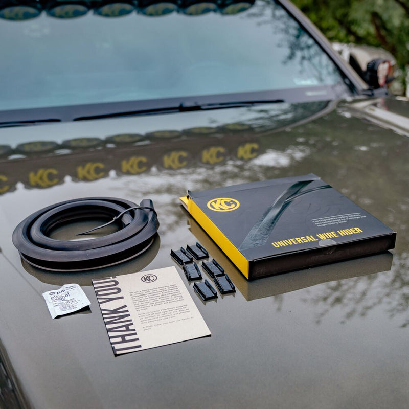 KC HiLiTES Universal Wire Hider with KClips on a vehicle: wire hider coil, six clips, an alcohol swab, a thank you card and a KC Universal Wire Hider box on a truck hood