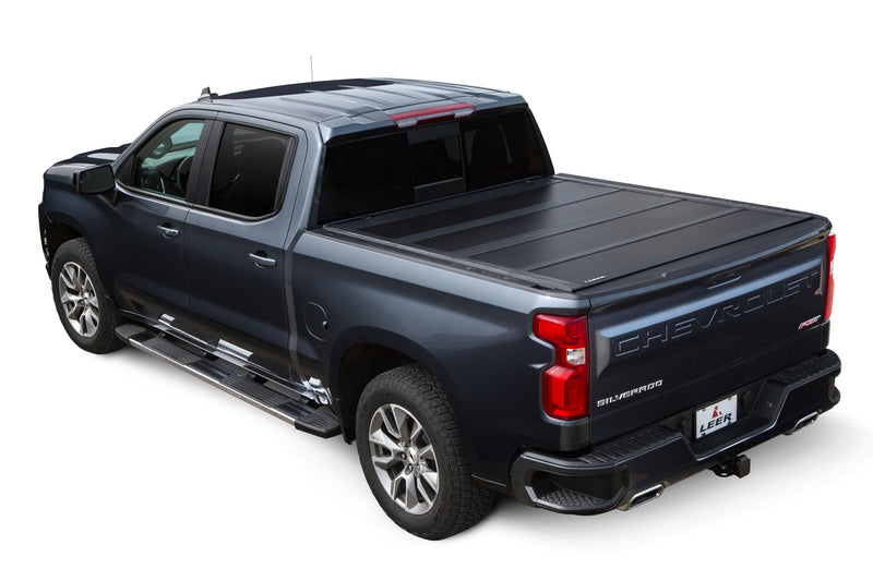 LEER HF650M hard folding tonneau cover on a pickup bed: black hard quad-fold cover closed over a dark blue pickup bed, rear three-quarter view (tow hitch accessory shown, not included)