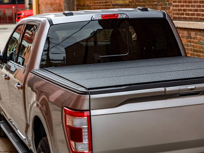Rear three-quarter view of a pickup bed with a diamond-pattern black hard tri-fold tonneau cover closed, brick wall behind