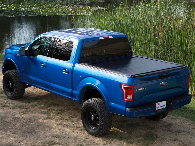 Pace Edwards BedLocker: black retractable cover fully closed on a lifted blue pickup parked by a pond
