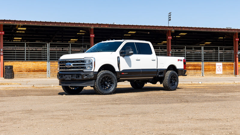 ReadyLIFT Coilover Front Lift Kit installed on a Ford F-250 Super Duty: white Ford Super Duty crew cab on black wheels in front of livestock pens under blue sky