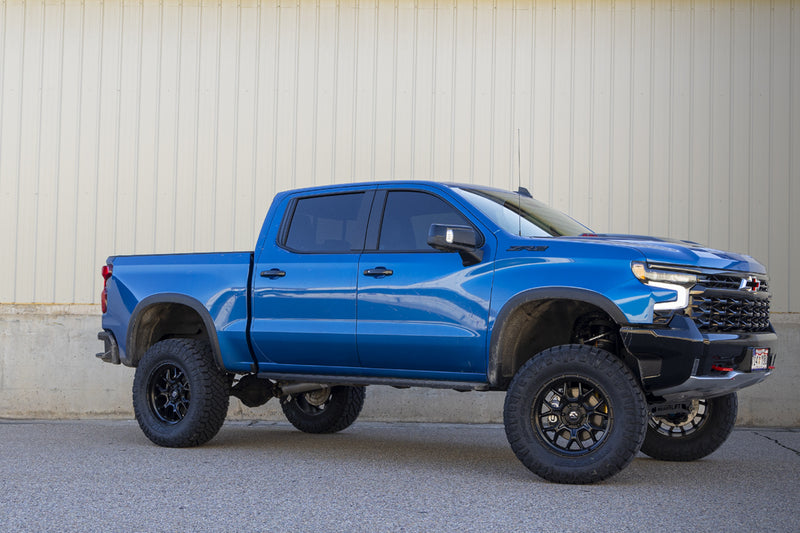 ReadyLIFT Lift Kit installed on a Chevrolet Silverado 1500 ZR2: blue Chevrolet Silverado ZR2 crew cab lifted on black wheels and all-terrain tires beside a concrete wall