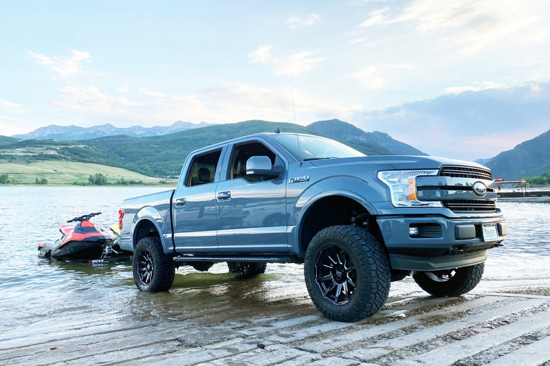 ReadyLIFT MAX3.3 Suspension System installed on a Ford F-150: blue Ford F-150 crew cab lifted on black wheels backing a personal watercraft down a lake ramp