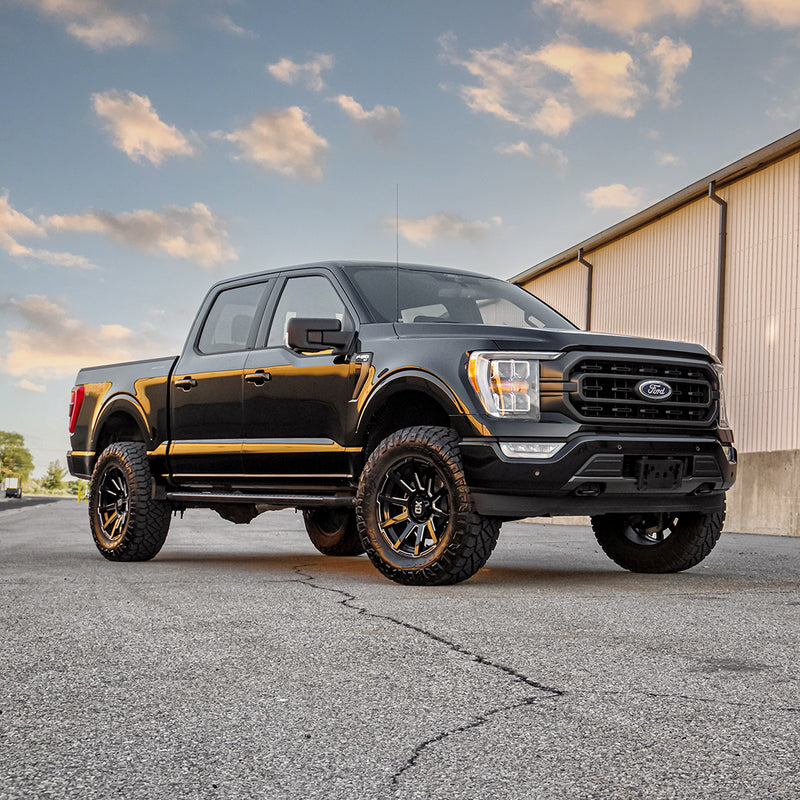ReadyLIFT SST Lift Kit installed on a Ford F-150: A black lifted Ford F-150 SuperCrew parked on pavement beside a building at sunset