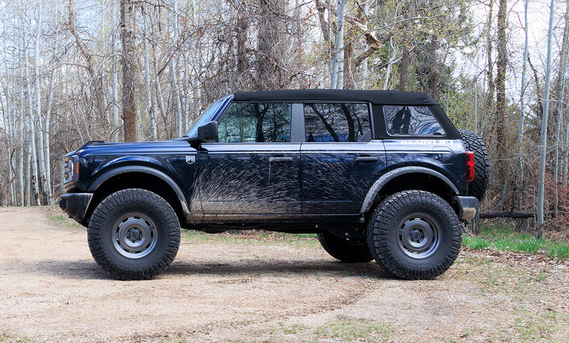 ReadyLIFT SST Lift Kit installed on a Ford Bronco: A dark blue lifted Ford Bronco four-door with a soft top parked on gravel in front of trees, side view