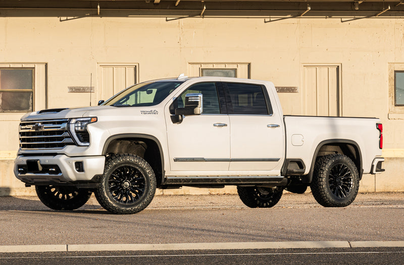 ReadyLIFT SST Lift Kit installed on a Chevrolet Silverado 2500HD: white Chevrolet Silverado HD High Country crew cab leveled on black wheels in front of a tan building