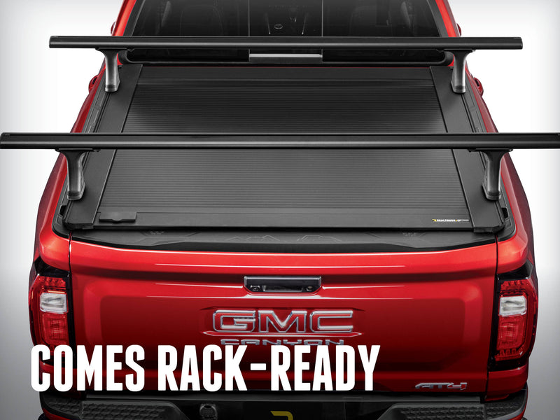 Rear view of a red pickup with the RetraxONE XR closed under two crossbars mounted in its T-slot rails, overlay headline comes rack-ready (crossbars sold separately)