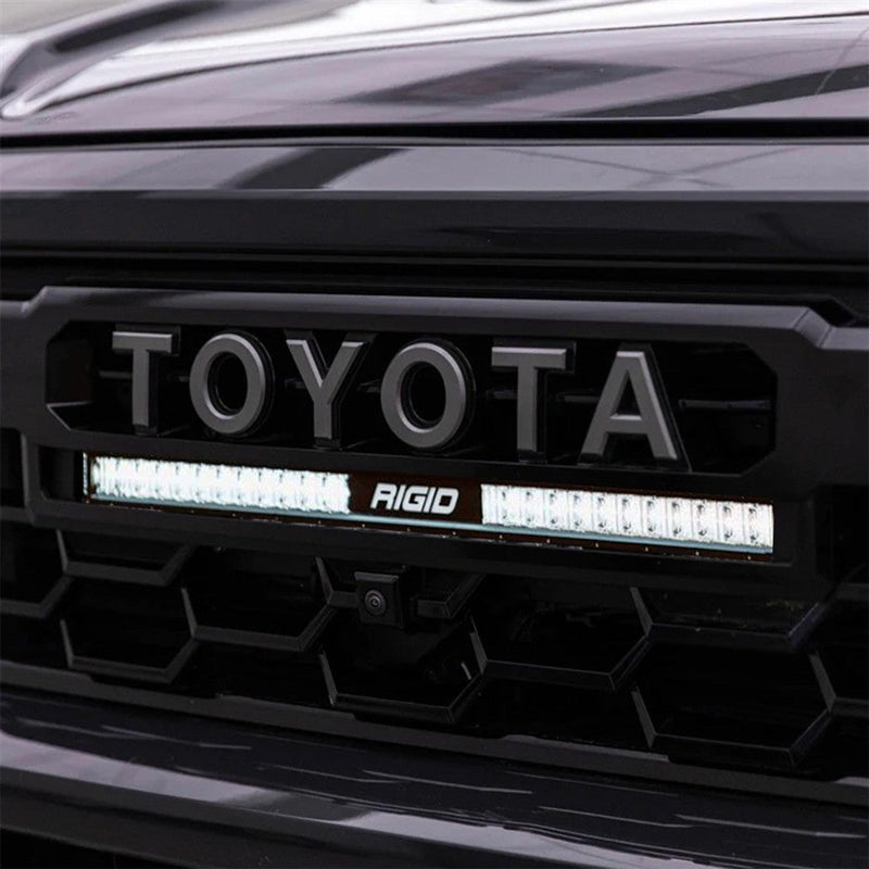 Rigid 20" SR-Series SAE Grille Kit installed on a Toyota Tacoma: close-up of a lit 20" SR-Series light bar installed under the TOYOTA lettering in a black Tacoma grille