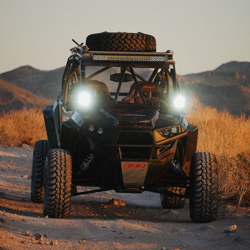 Rigid Adapt XP Pair installed on a Polaris RZR: A side-by-side UTV on a desert track at sunset with two white Adapt XP lights shining from its A-pillars